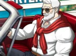 Hombre musculoso con barba blanca y gafas oscuras sentado en un descapotable rojo vistiendo camisa blanca y suéter rojo.