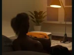 Mujer rubia desnuda recostada en cama mostrando sus pechos mientras acaricia a alguien en una habitación iluminada.